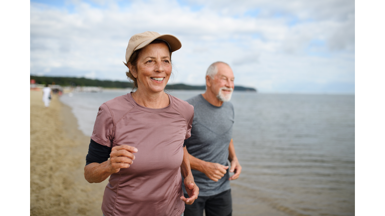 Active senior couple runners jogging outdoors on sandy beach by sea in early morning.