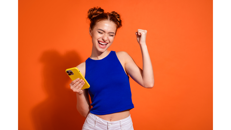 Excited Young Woman with Stylish Hairstyle Celebrating Success Holding Smartphone Against Vibrant Orange Background