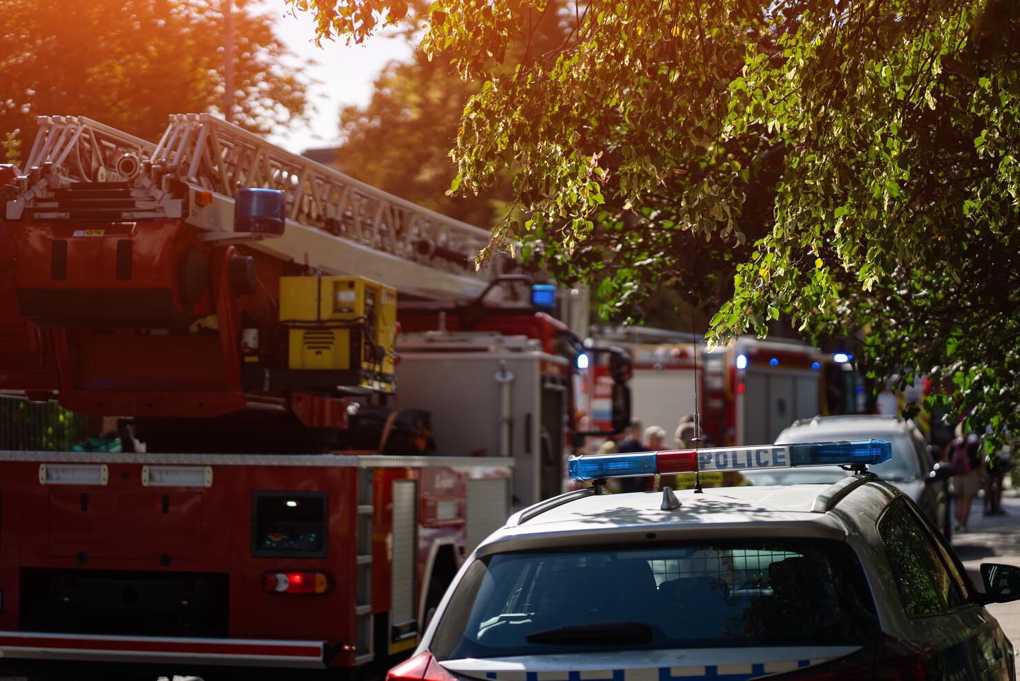Firefighters and police respond to emergency, fire trucks with ladders and flashing blue lights. Firemen in gear prepare equipment in residential area