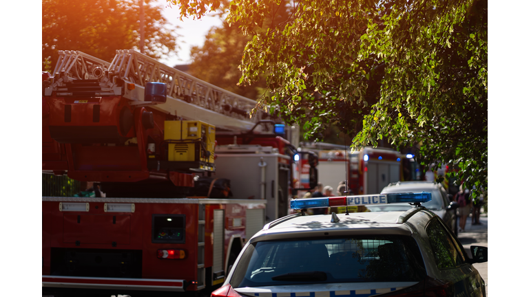 Firefighters and police respond to emergency, fire trucks with ladders and flashing blue lights. Firemen in gear prepare equipment in residential area