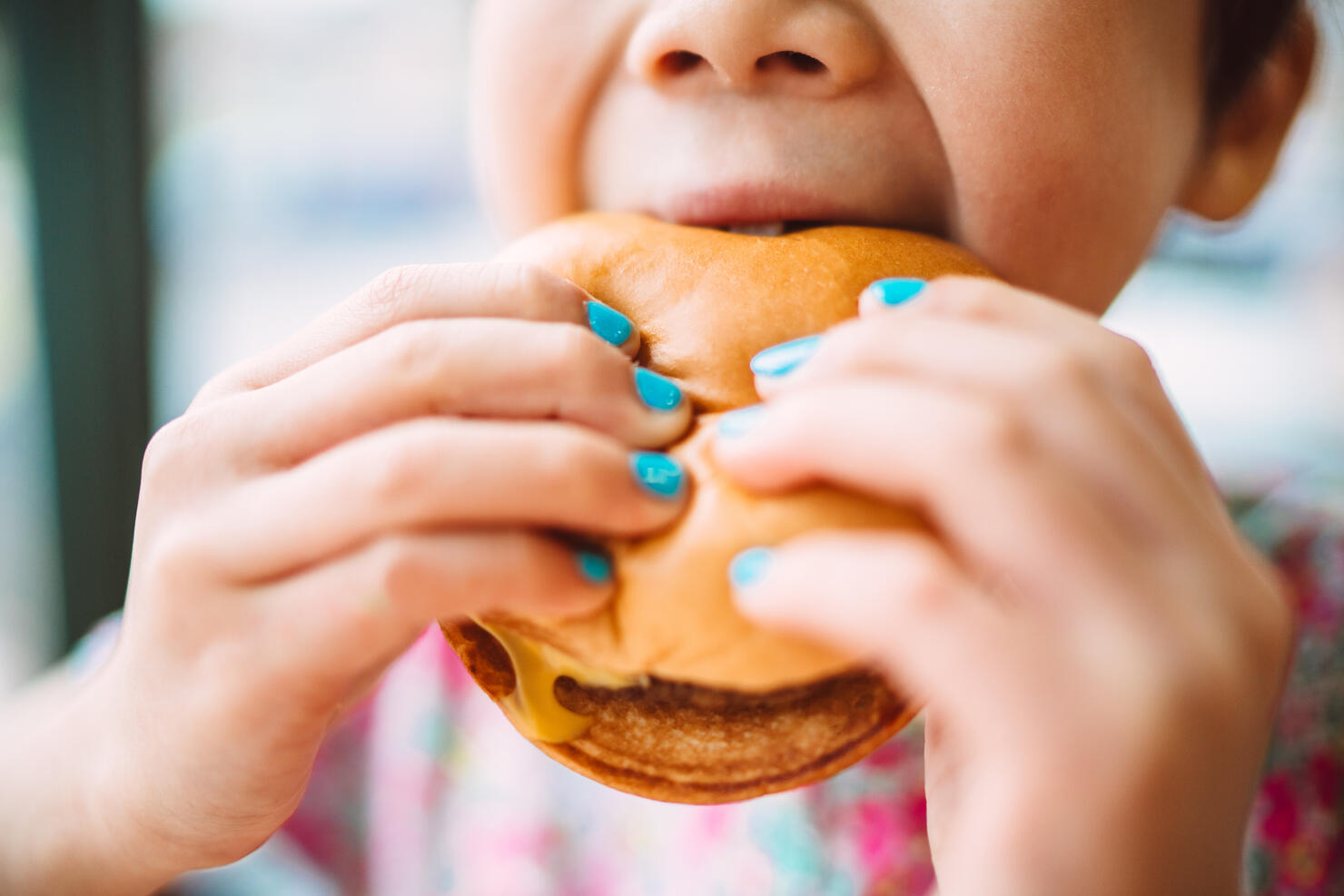 Close-up of child enjoying a cheeseburger with brightly painted nails