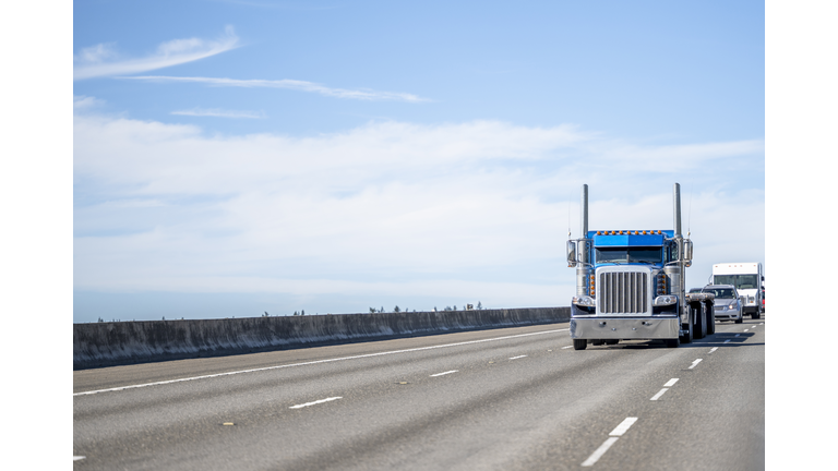 Stylish blue big rig semi truck tractor with empty flat bed semi trailer running on the wide highway road in front of another traffic