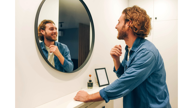 Handsome bearded guy looks at himself in the mirror. Jeans shirt. Brod guy. Admires himself. Nice appearance. Long hair. Casual style.