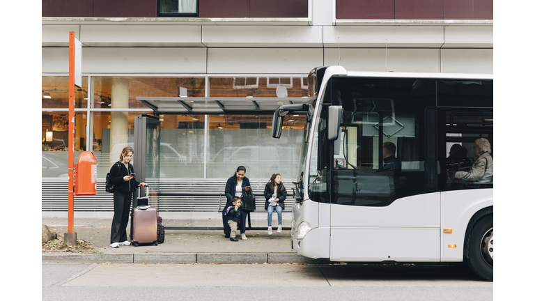 Full length of woman with luggage and family waiting for bus at bus stop in city