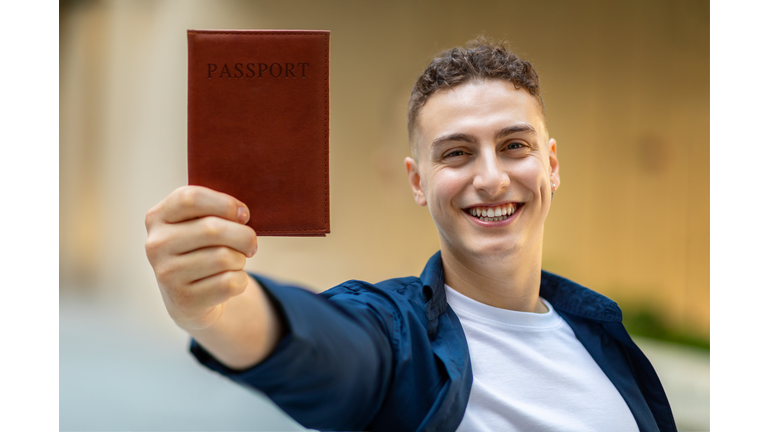 A joyful young man holding up a passport with a brown cover towards the camera
