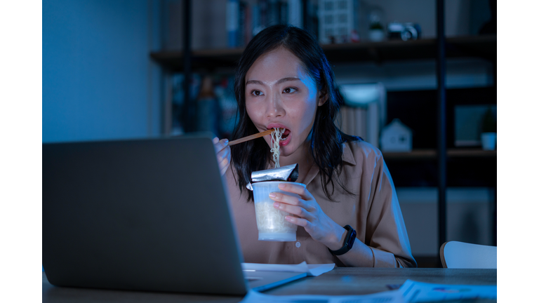 Asia freelance smart business women eating instant noodles while working on laptop in living room at home at night