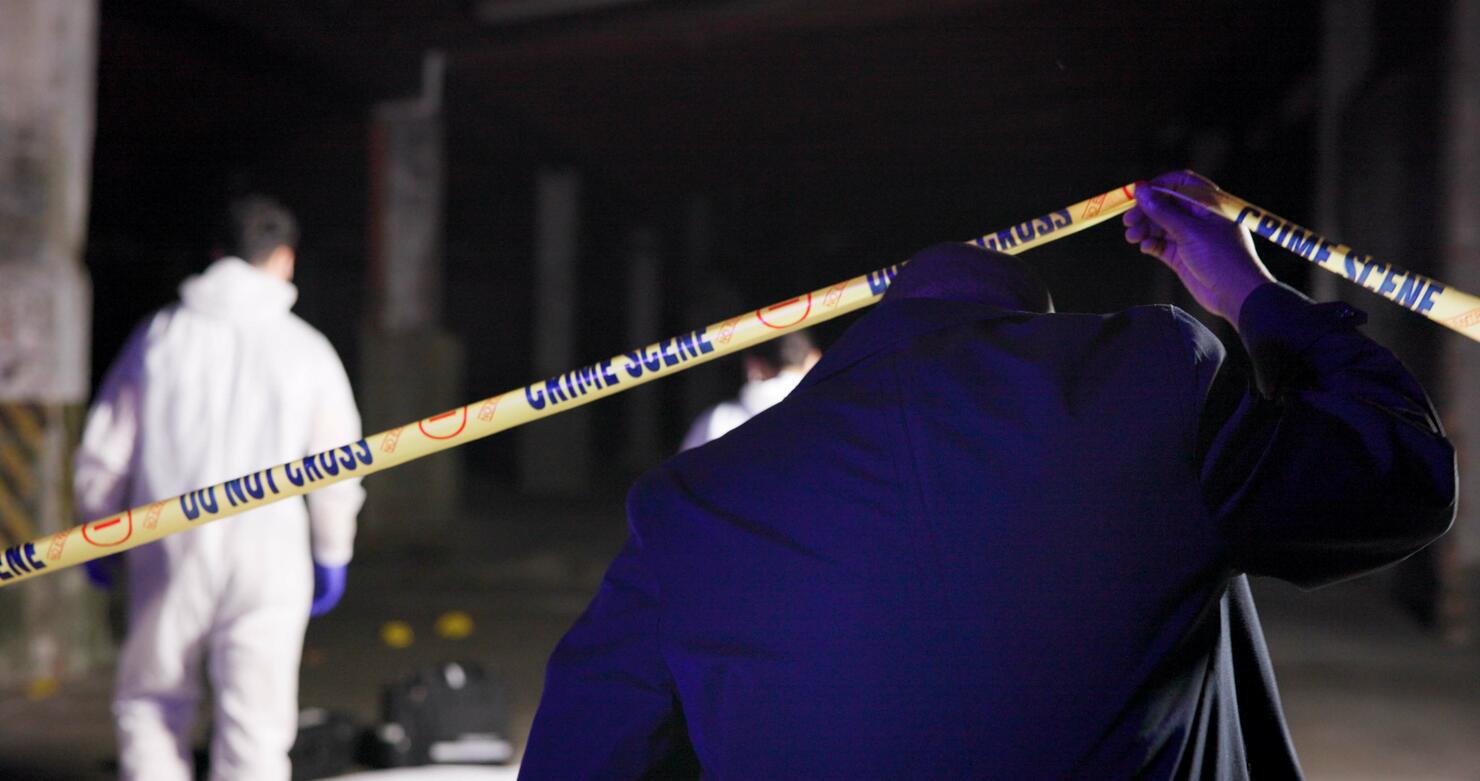 Detective, yellow tape and back at crime scene with forensic team, inspection and outdoor at night. Police, csi and barrier for investigation, evidence and search for lead, emergency and law in city