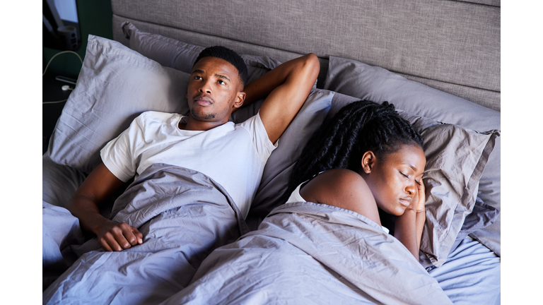Young man looking unhappy while lying in bed with his sleeping wife