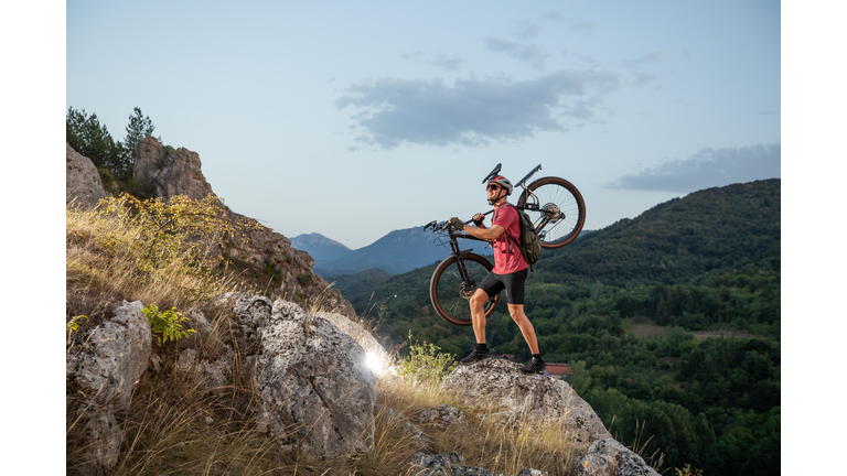 Cyclist Carrying Mountain Bike on Rocky Path in Wilderness at Sunrise