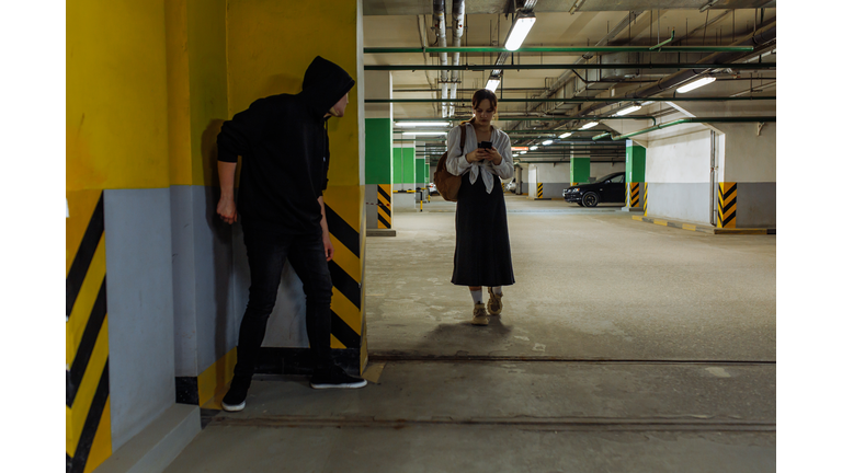 Unsuspicious teenage girl walking in underground parking lot using smart phone while male stalker waiting to attack