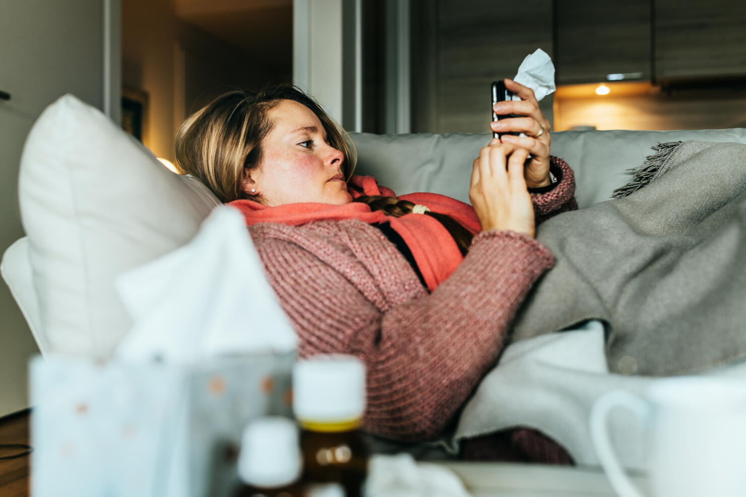 Woman lying on a sofa and using her mobile phone writing a sick note while having a flu and feeling sick.