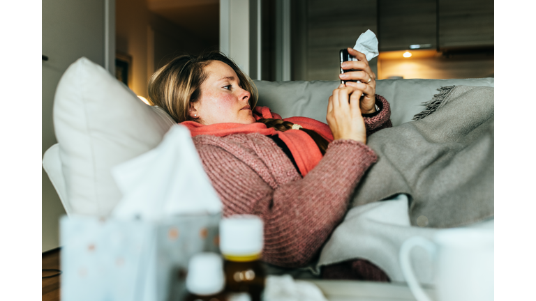 Woman lying on a sofa and using her mobile phone writing a sick note while having a flu and feeling sick.