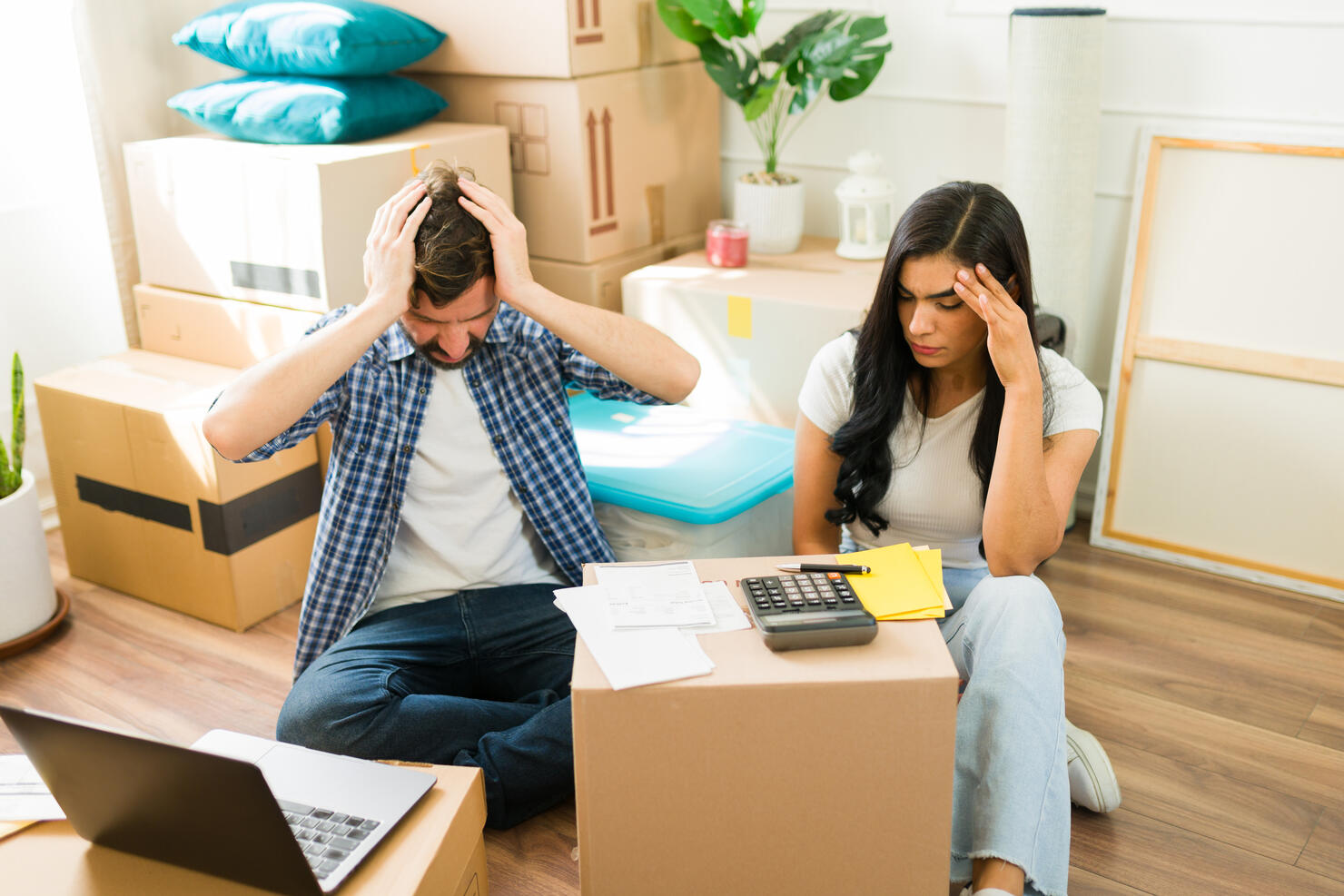 Young Hispanic couple surrounded by moving boxes in their new home, feeling overwhelmed while working on their budget