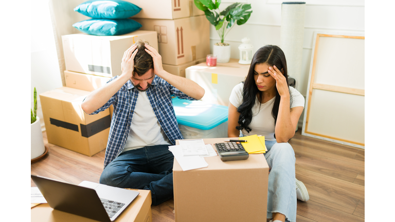 Young Hispanic couple surrounded by moving boxes in their new home, feeling overwhelmed while working on their budget