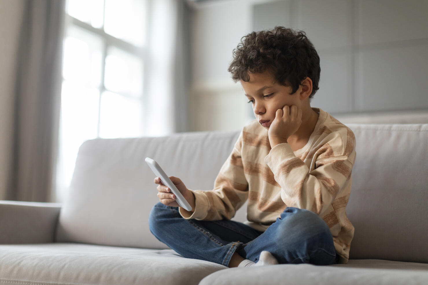 Bored black preteen boy sitting on couch and using smartphone at home