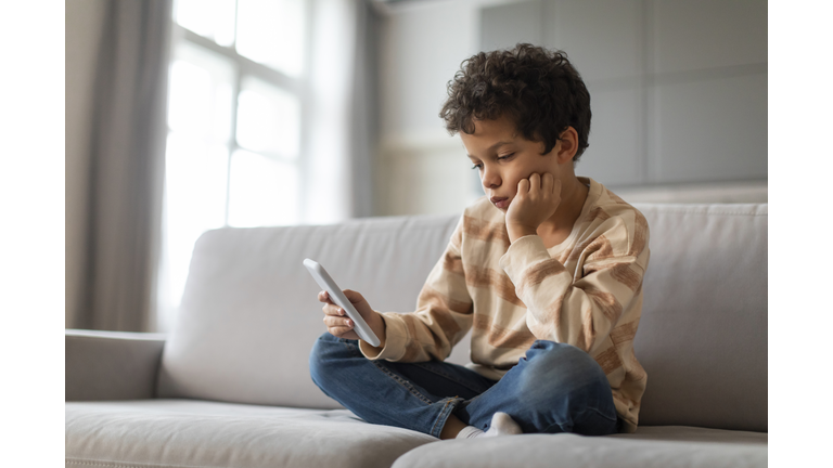 Bored black preteen boy sitting on couch and using smartphone at home