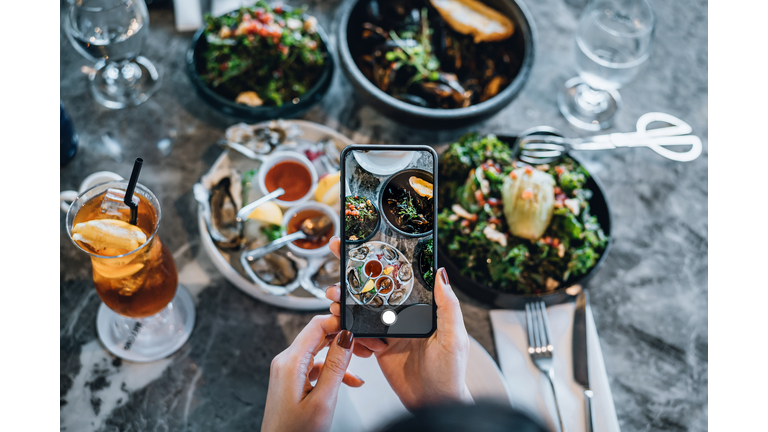Overhead view of young woman taking photos of scrumptious and delicious meal on dining table with smartphone before eating it in restaurant. Eating out lifestyle. Camera eats first culture