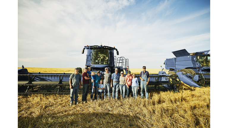 Wide shot portrait of smiling multigenerational family standing in wheat field in front of combines during summer harvest