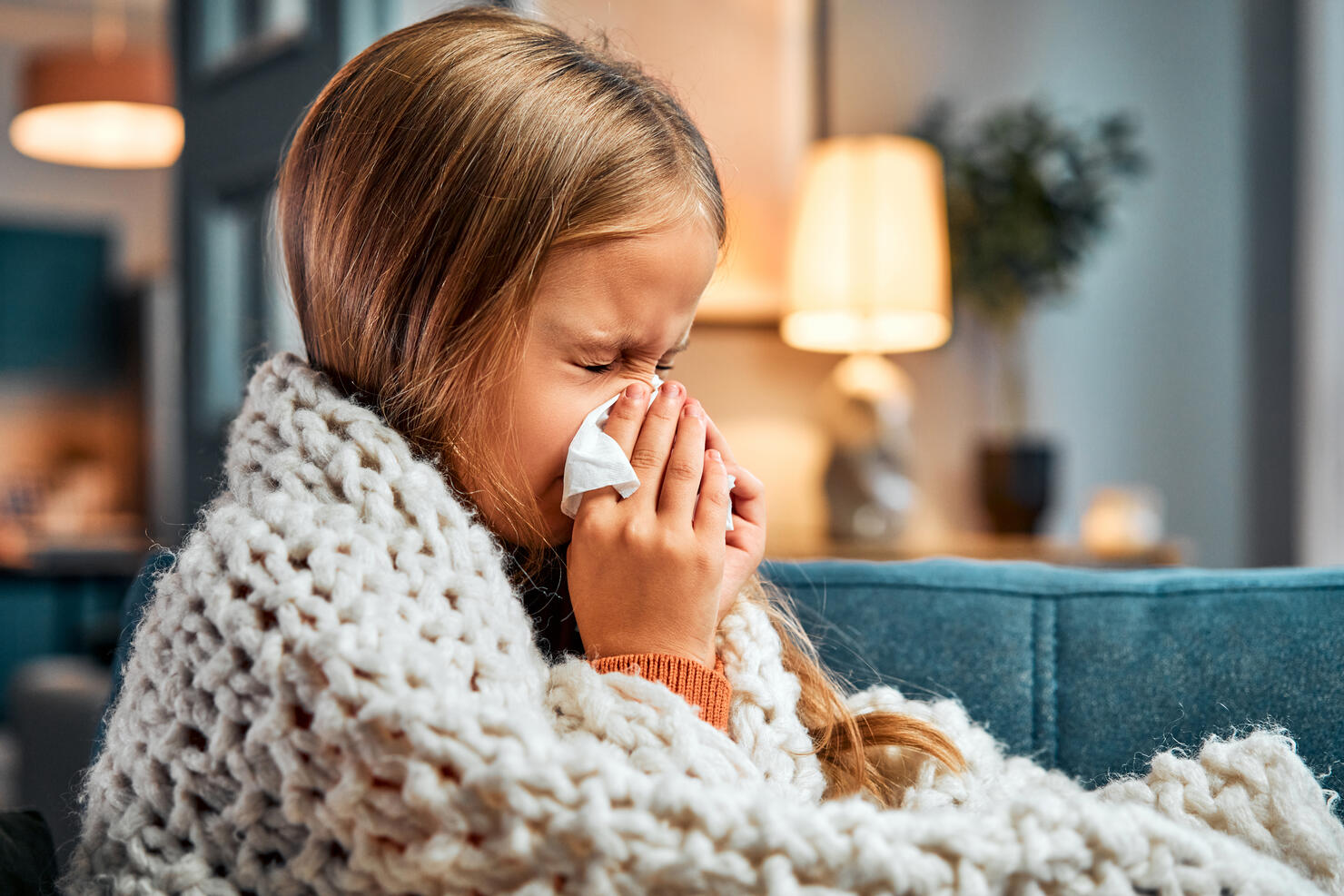 Season of flu. Little kid covered with blanket pressing handkerchief to nose while sneezing at apartment