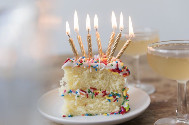 Candles burning on slice of cake with sprinkles near champagne