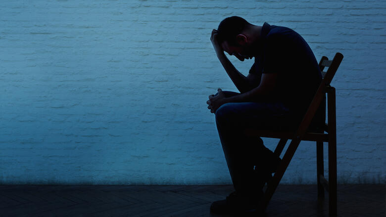 Unhappy and lonely young man in front of a wall. He sits on a chair and thinks alone under blue light. Negative human expression.