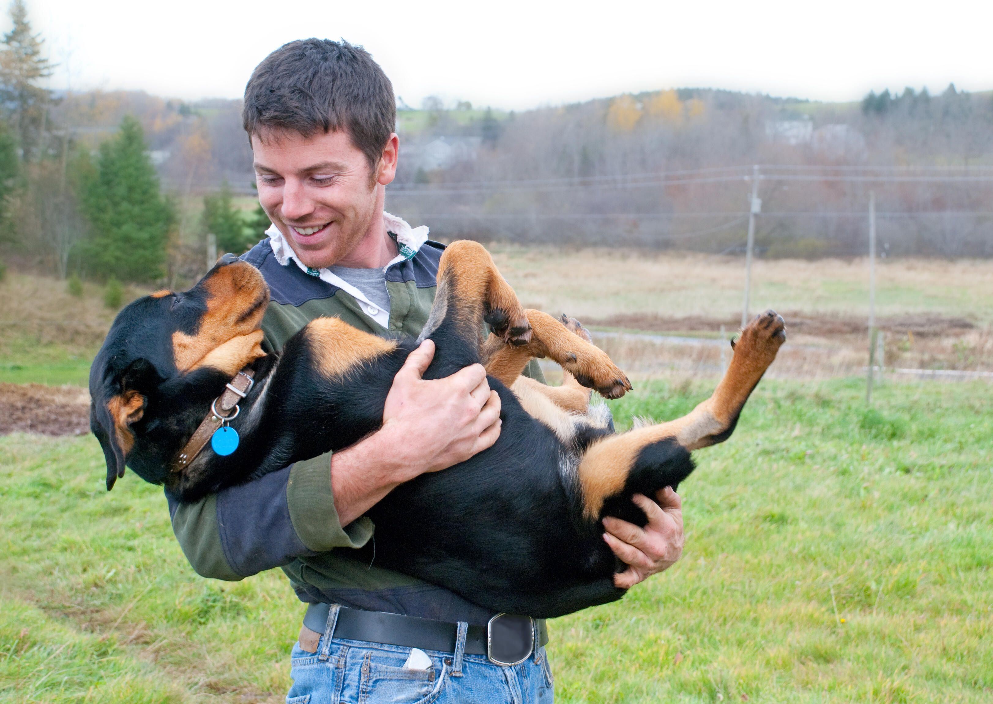 Handsome smiling young man lifts and cradles a half grown Rottweiler puppy, which is relaxed and looking at the man adoringly