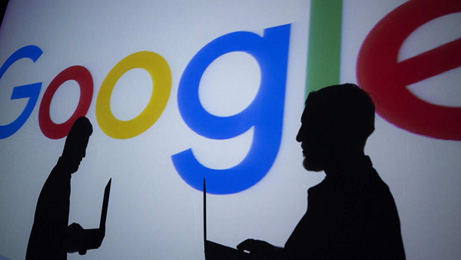 Silhouettes of people holding laptops are seen in front of the logo of 'Google' technology company on the 20th anniversary of Google