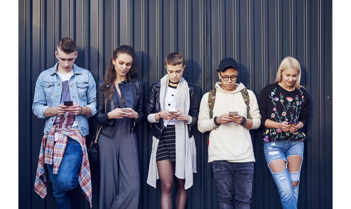 Row of five young adult friends leaning against black wall looking at smartphones