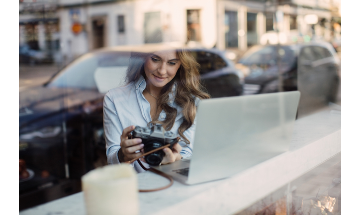 Young woman holding camera while sitting with laptop at cafe seen through window glass