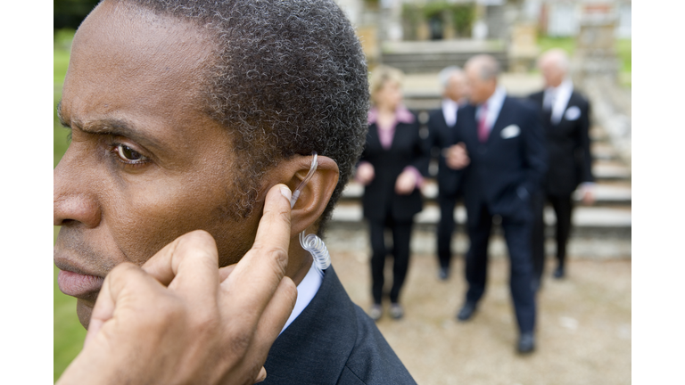 Man with hand on earpiece by manor house, businessmen and women in background, close-up