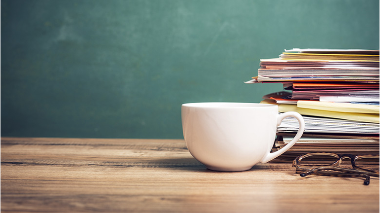 Coffee cup, papers stack on wooden school desk with chalkboard.