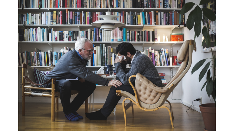 Senior therapist counseling young patient while sitting by bookshelf during therapy session