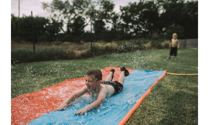 Boy sliding on water slide at yard with brother standing in background