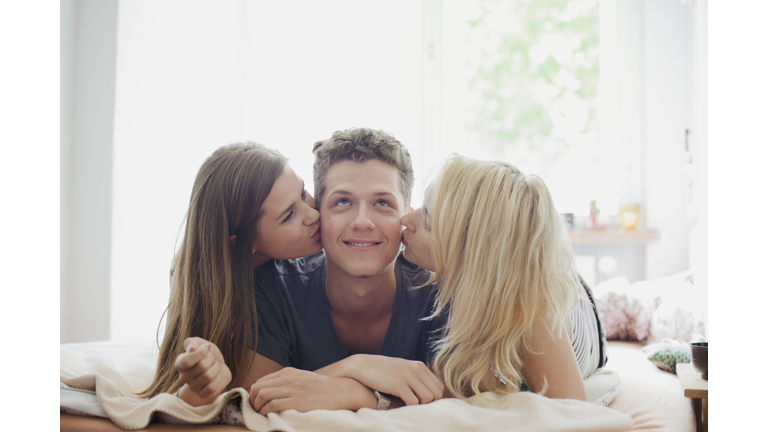 Young female friends kissing handsome man lying on bed at home