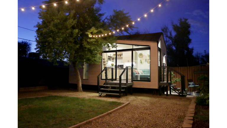 A tiny house with large glass windows, sits in the backyard at night, surrounded by trees and party lights.