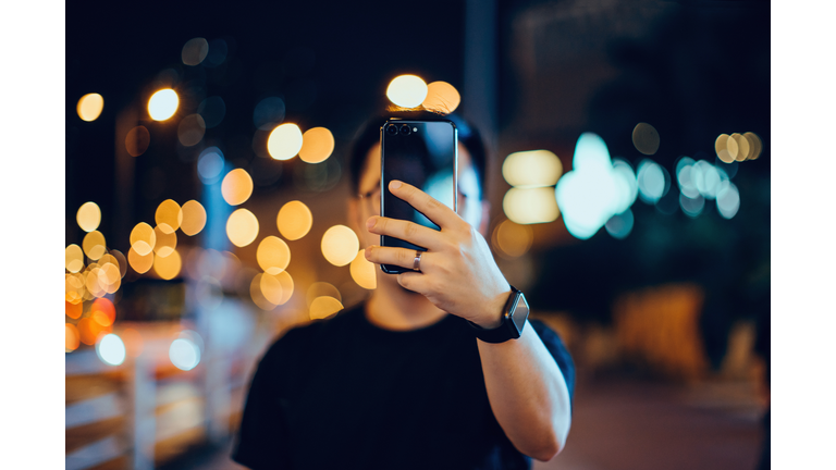 Smart young Asian man taking pictures with smartphone in city street, against illuminated city street light and city traffic