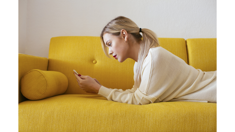 Blonde woman lying on sofa, using smartphone at home