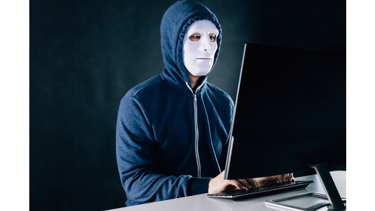 Anonymous and masked hacker under hoodie using computer isolated over dark background - illegal online internet criminal concept.