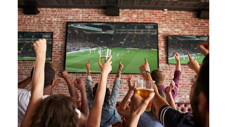 Rear View Of Friends Watching Game In Sports Bar On Screens