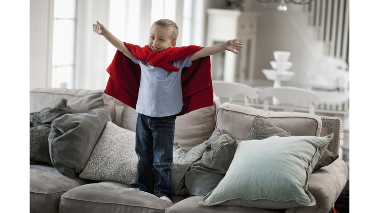 A child standing wearing a red cape, arms raised in a superhero pose. 