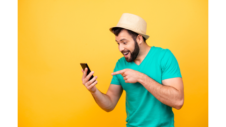 Ha-ha! So funny! Portrait of happy excited crazy man with stubble wearing hat and green tshirt, he is pointing on his smartphone and rejoicing