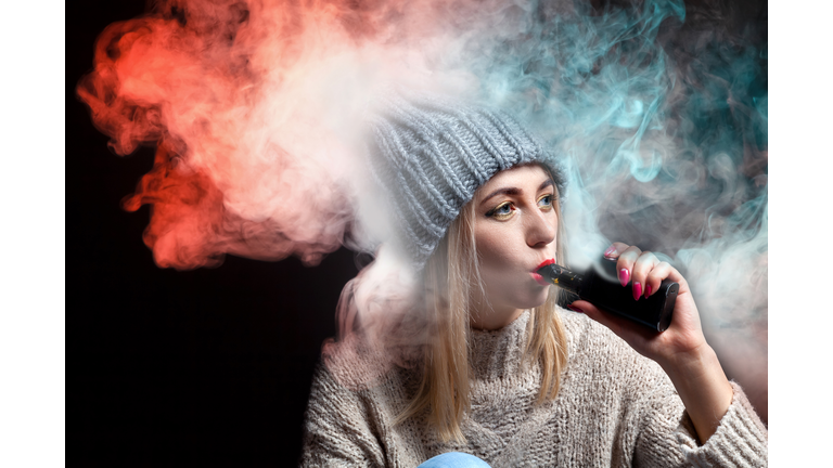 Blonde girl in a knitted sweater and hat holds an electronic cigarette vape in her hand with a manicure and exhales a lot of colored red and green smoke, with red lipstick on a black background.