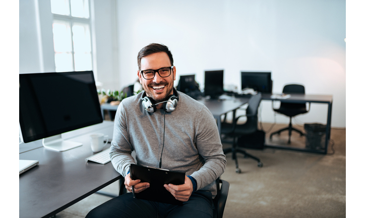 Portrait of a smiling young man with headset sitting in front of computer, looking at camera, in modern office.