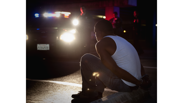 Police shining lights on handcuffed African man sitting on curb