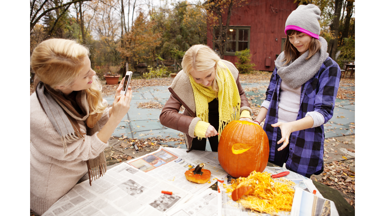 Woman photographing while friends carving Halloween pumpkin at table