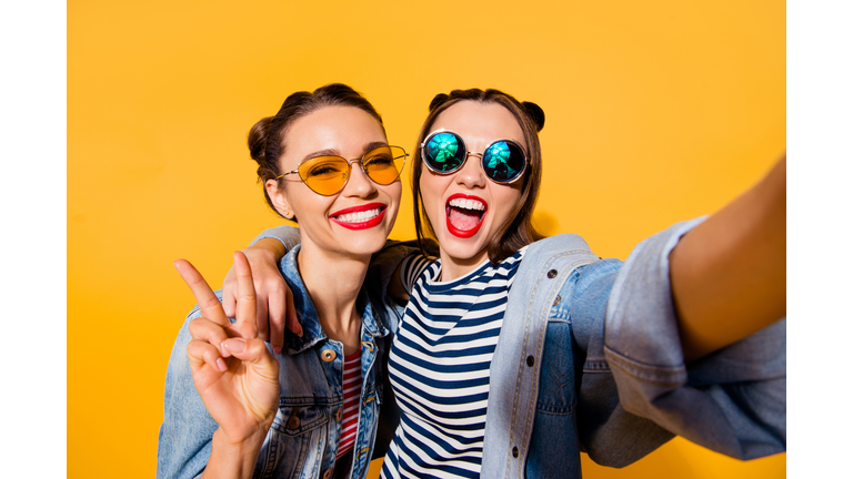 Two glad positive grinning lady stand in glasses spectacles street style stylish trendy cool casual denim jeans clothes isolated on yellow background in take picture on cellular make hollywood smile