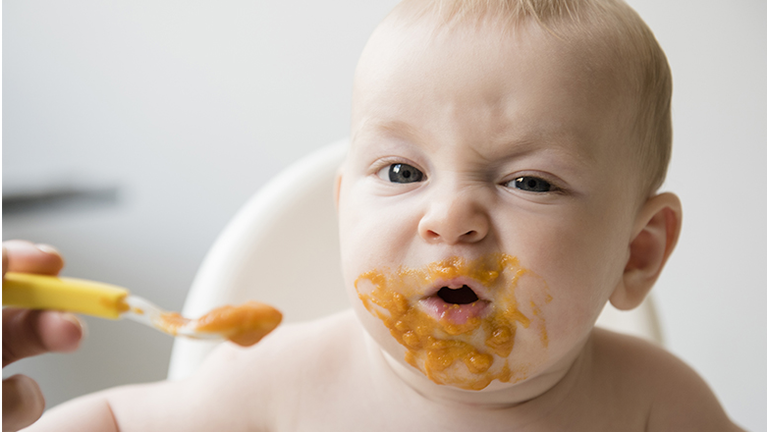 Mother feeding messy baby son with spoon in high chair