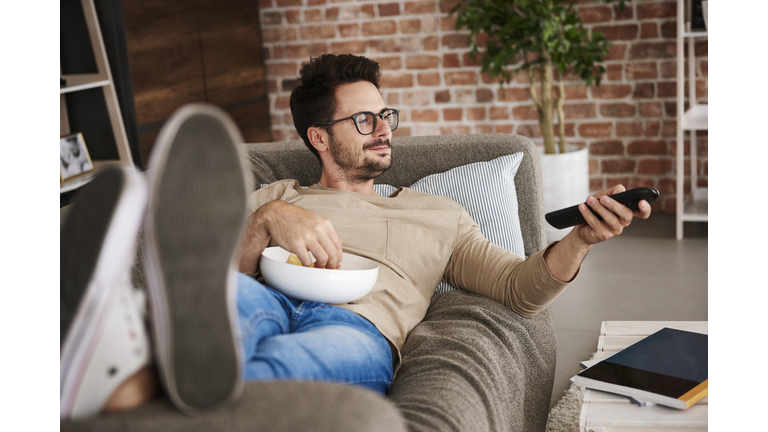 Content man lying on couch at home with bowl of potato chips watching TV