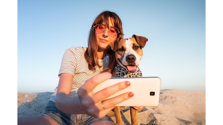 Low Angle View Of Young Woman Taking Selfie With Dog Through Smart Phone At Beach Against Clear Sky