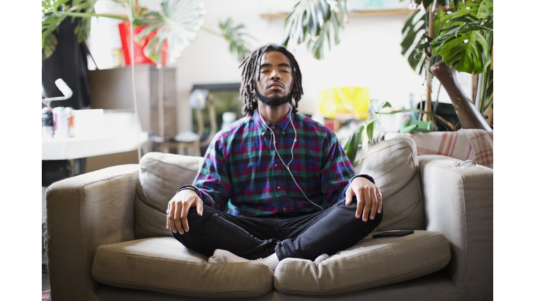 Serene young man meditating with headphones on apartment sofa